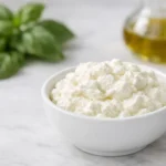 Fromage ricotta frais et onctueux dans un bol blanc, avec des herbes et de l'huile d'olive en arrière-plan flou.