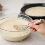 Pâte à crêpes lisse et prête à l'emploi dans un bol, avec une crêpe en cuisson en arrière-plan. Recette pâte à crêpes sans re